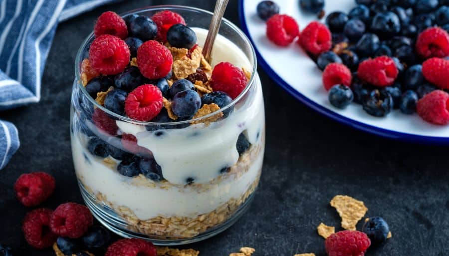 A glass filled with layered granola and yogurt and topped with fresh raspberries and blueberries.