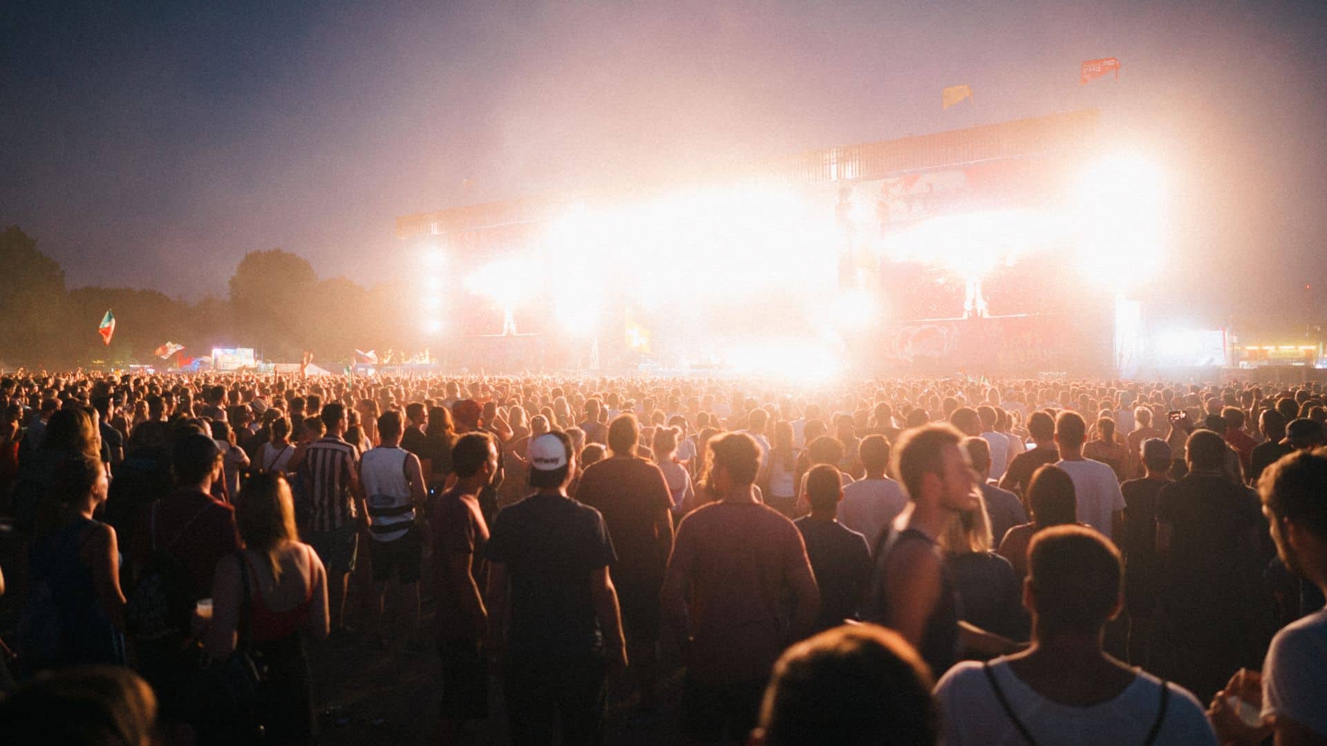 a large crowd of people at an outdoor music festival. the stage shines bright against the dark evening sky.