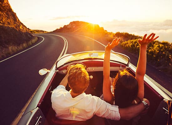 Couple driving along the California coast in a red convertible car with the sun shining ahead of them