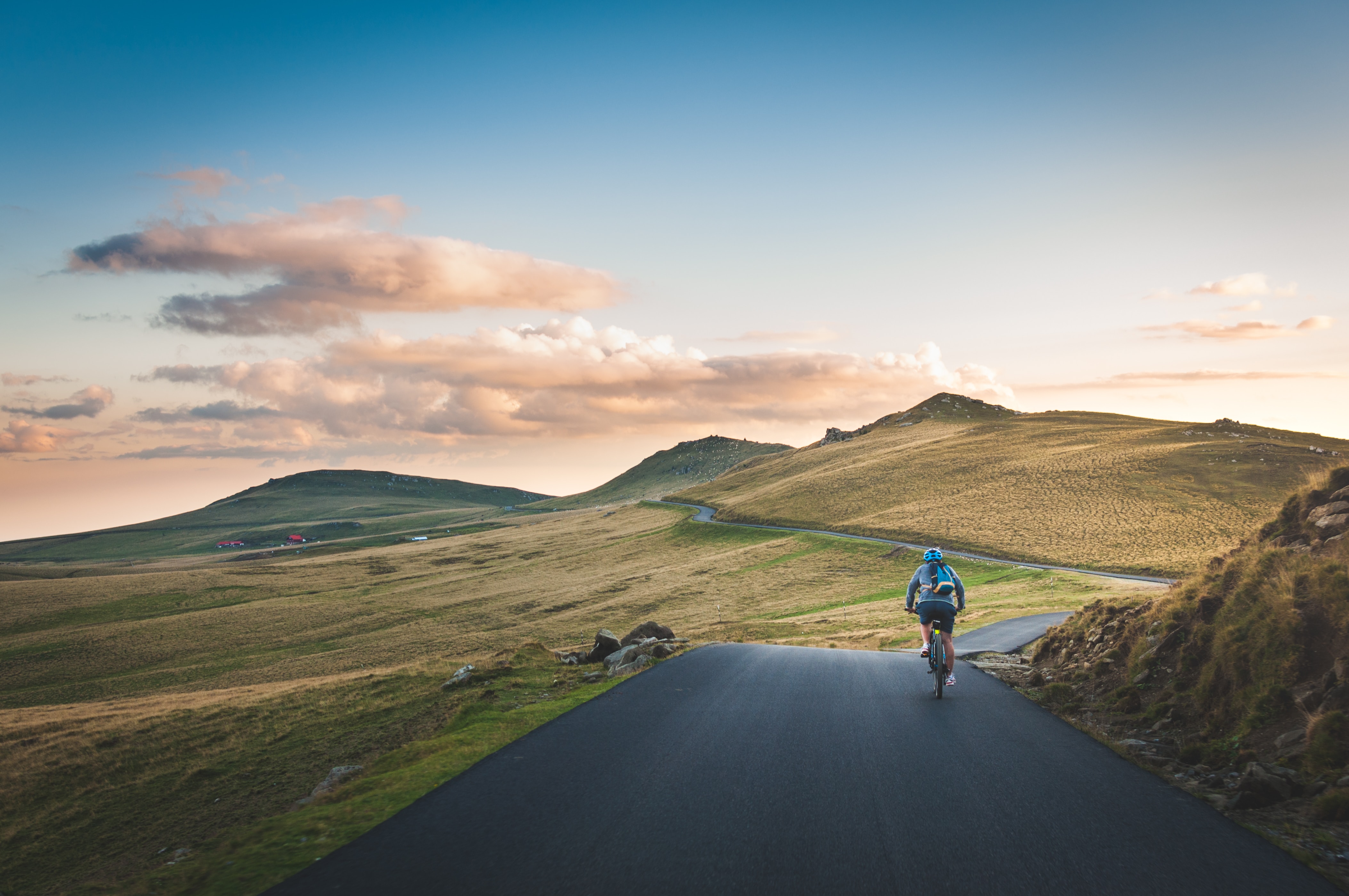 A cyclist is on a paved road that winds along the countryside. Light, evening clouds dot the landscape.