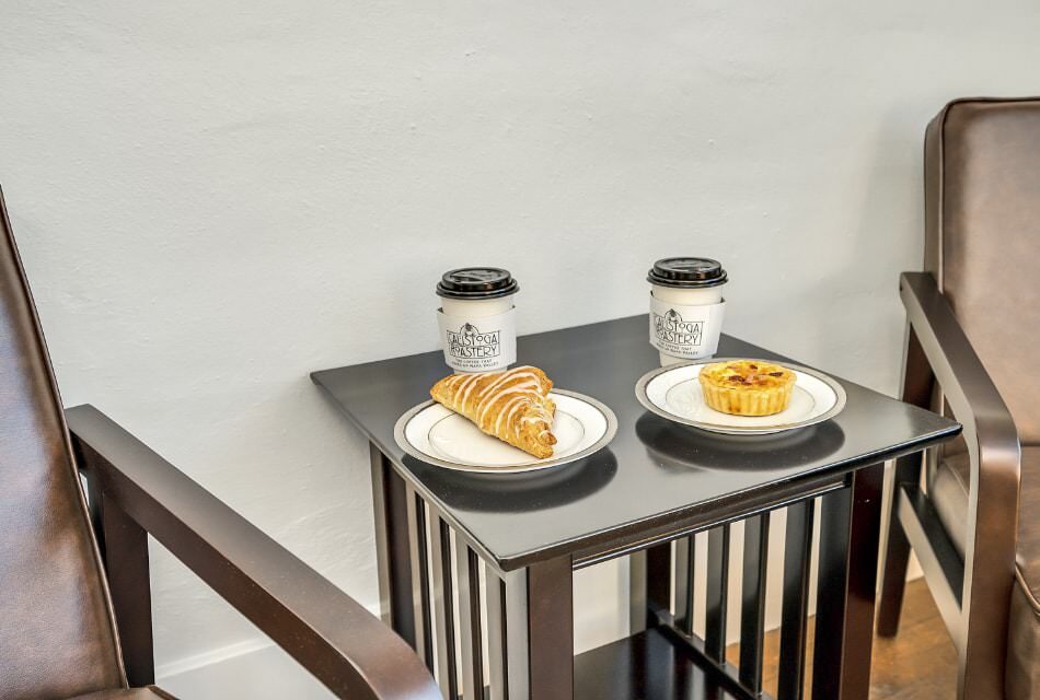 Pastry drizzled with white icing on a white plate and a small quiche on another white plate next to togo coffee cups on dark wooden end table