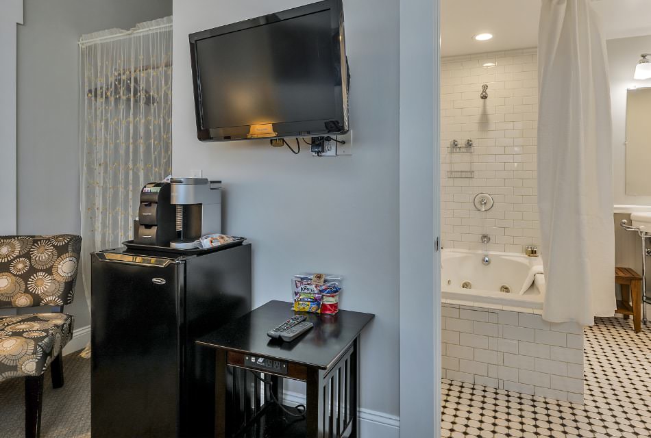 Bedroom with white walls, carpeting, mini fridge, wall-mounted flat-screen TV, and view into bathroom with white tiled tub