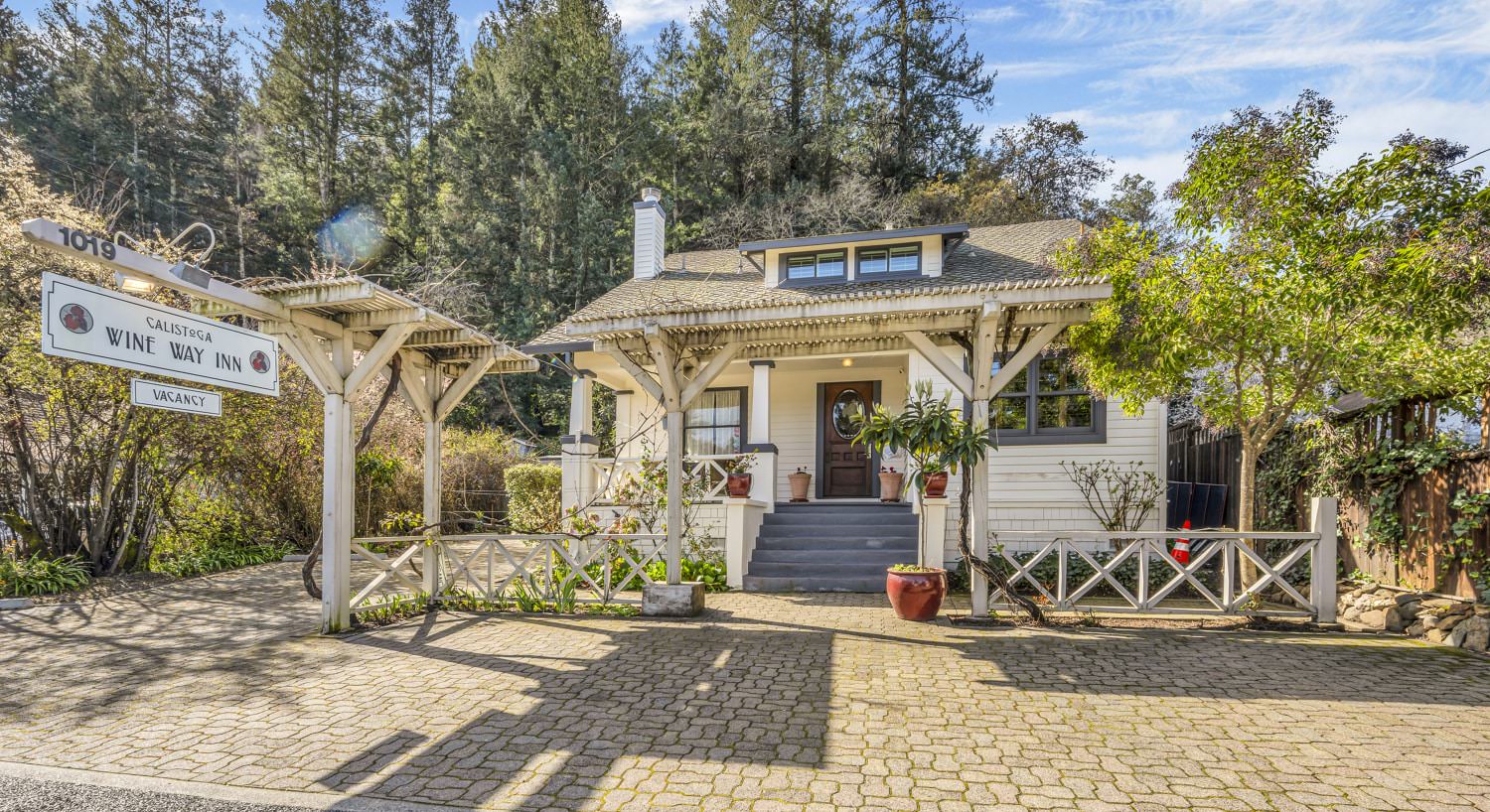 Front view of property painted white with gray trim, gray steps up to wooden door, and large trees in the background