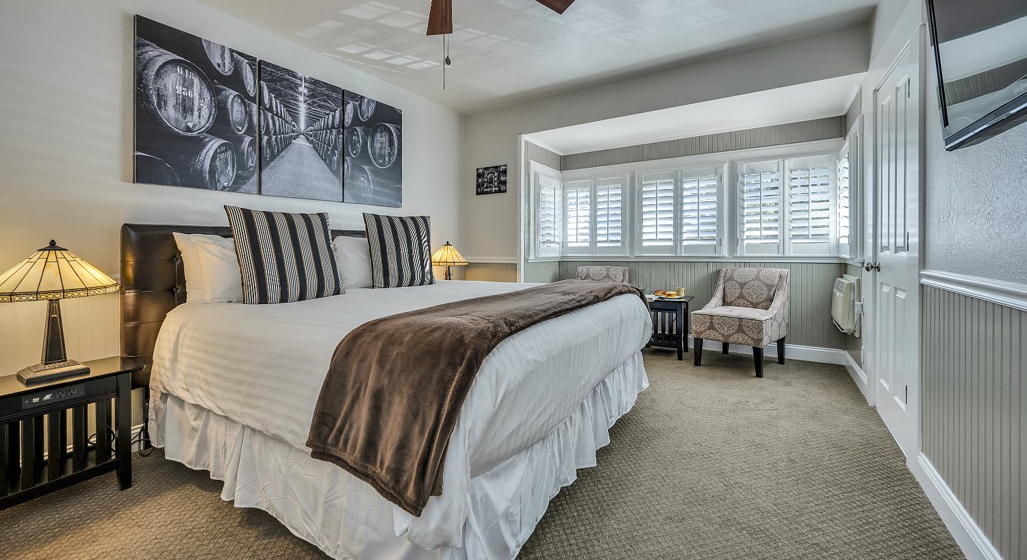 Bedroom with gray wainscotting, white walls, taupe carpeting, white bedding, and sitting area