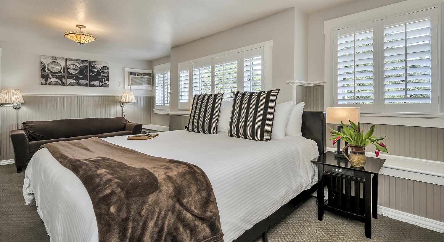 Bedroom with white walls, light gray wainscotting, light colored carpeting, white bedding, and upholstered brown sofa