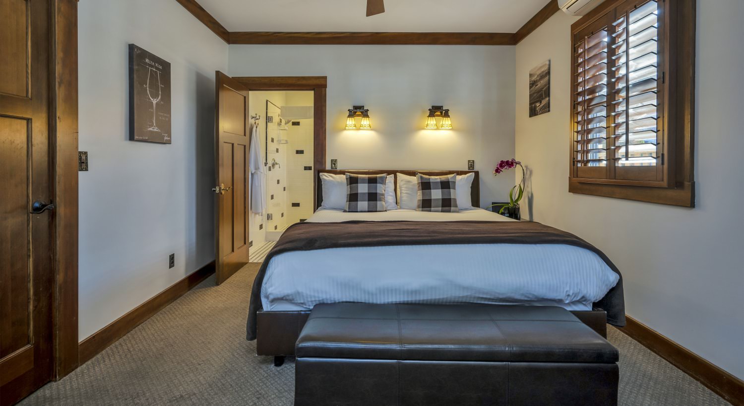 Bedroom with white walls, brown trim, light colored carpeting, white bedding, and upholstered leather bed bench