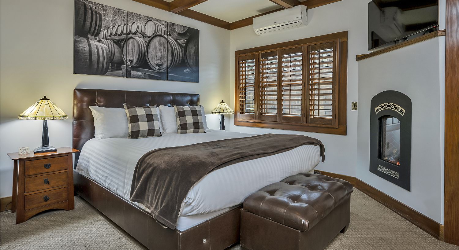 Bedroom with white walls, light colored carpeting, white bedding, leather bed bench, and electric fireplace