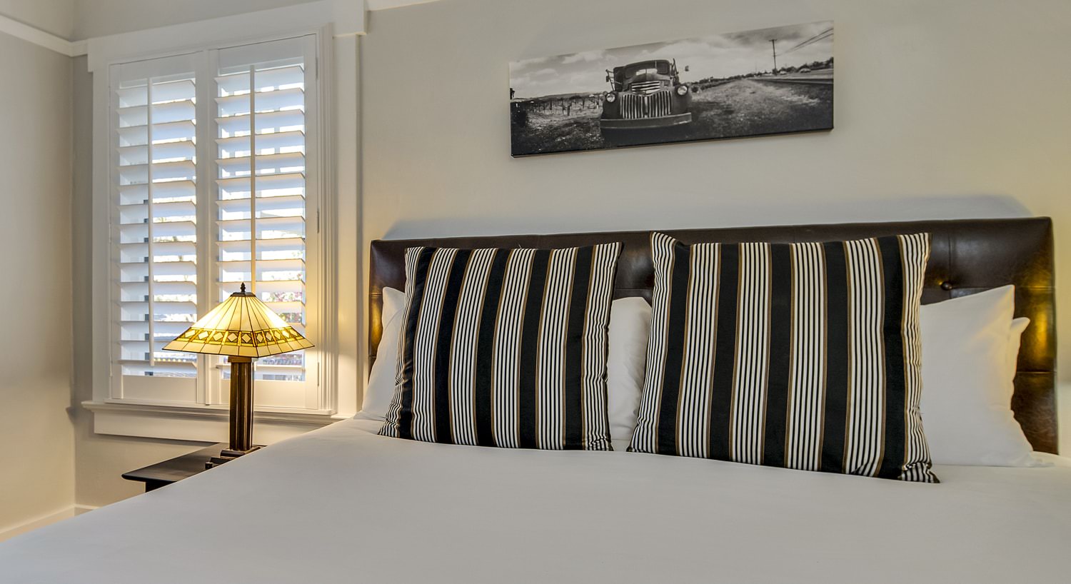 Bedroom with white walls, white trim, leather upholstered headboard, white bedding, and brown and white striped pillows