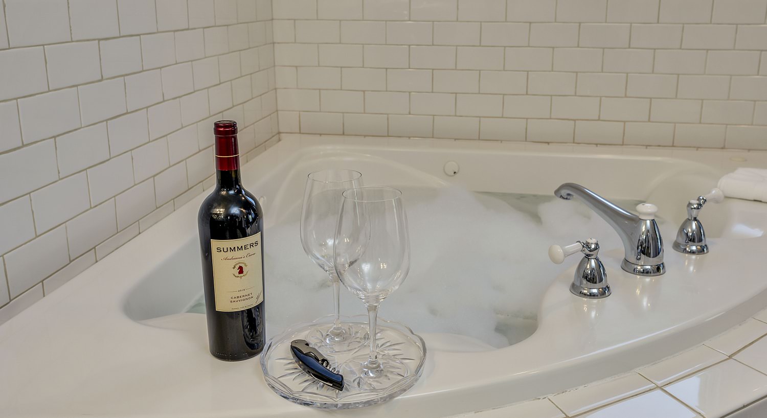 Two wine glasses on glass tray next to bottle of red wine on top of soaker tub surrounded by white tile