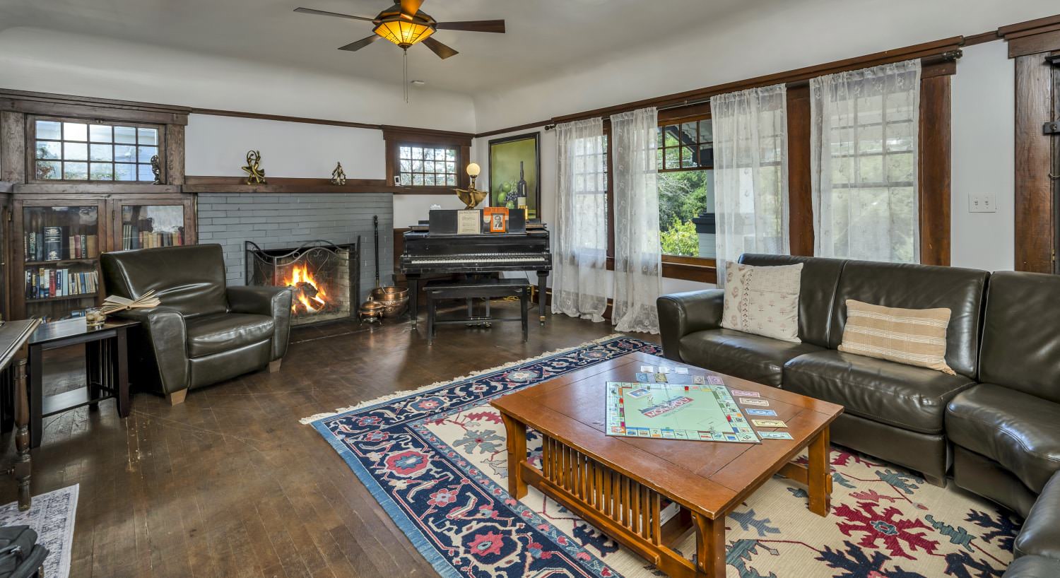 Large living room with white walls, brown wooden trim, hardwood flooring, leather sectional sofa, wooden coffee table, fireplace, and black piano