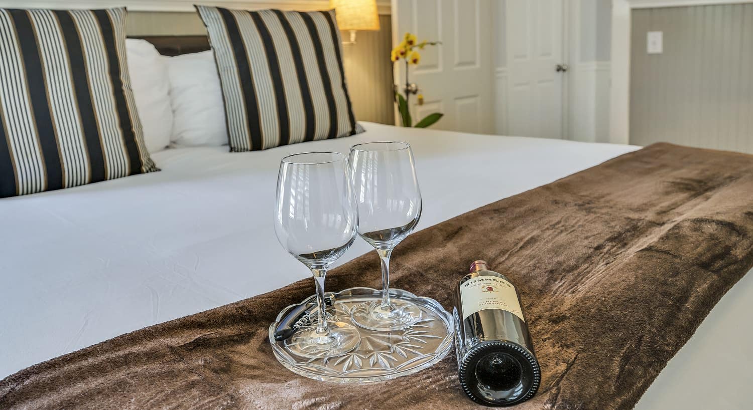 Close up view of two wine glasses on glass tray and bottle of wine on brown blanket on bed with white sheets
