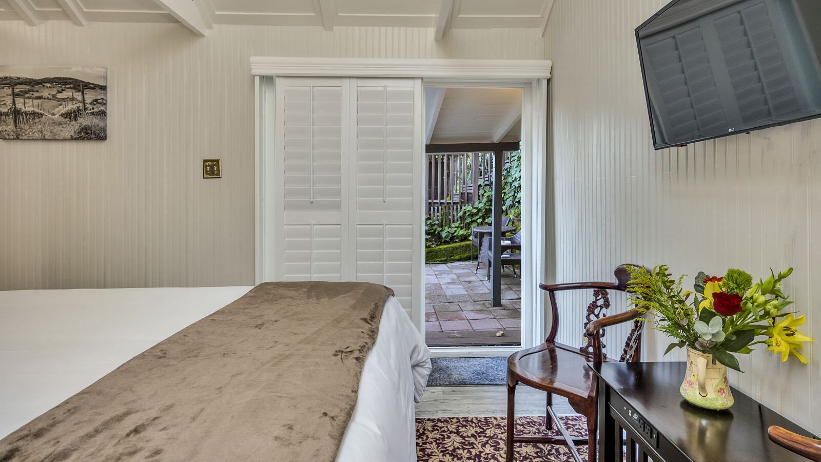 Bedroom with white walls, floral carpeting, white bedding, dark wooden table, and view out to back patio