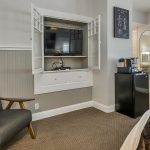 Bedroom with white walls, light gray wainscotting, light colored carpeting, white bedding, black mini fridge, and flatscreen TV