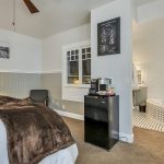 Bedroom with white walls, light gray wainscotting, light colored carpeting, white bedding, black mini fridge, and view into bathroom