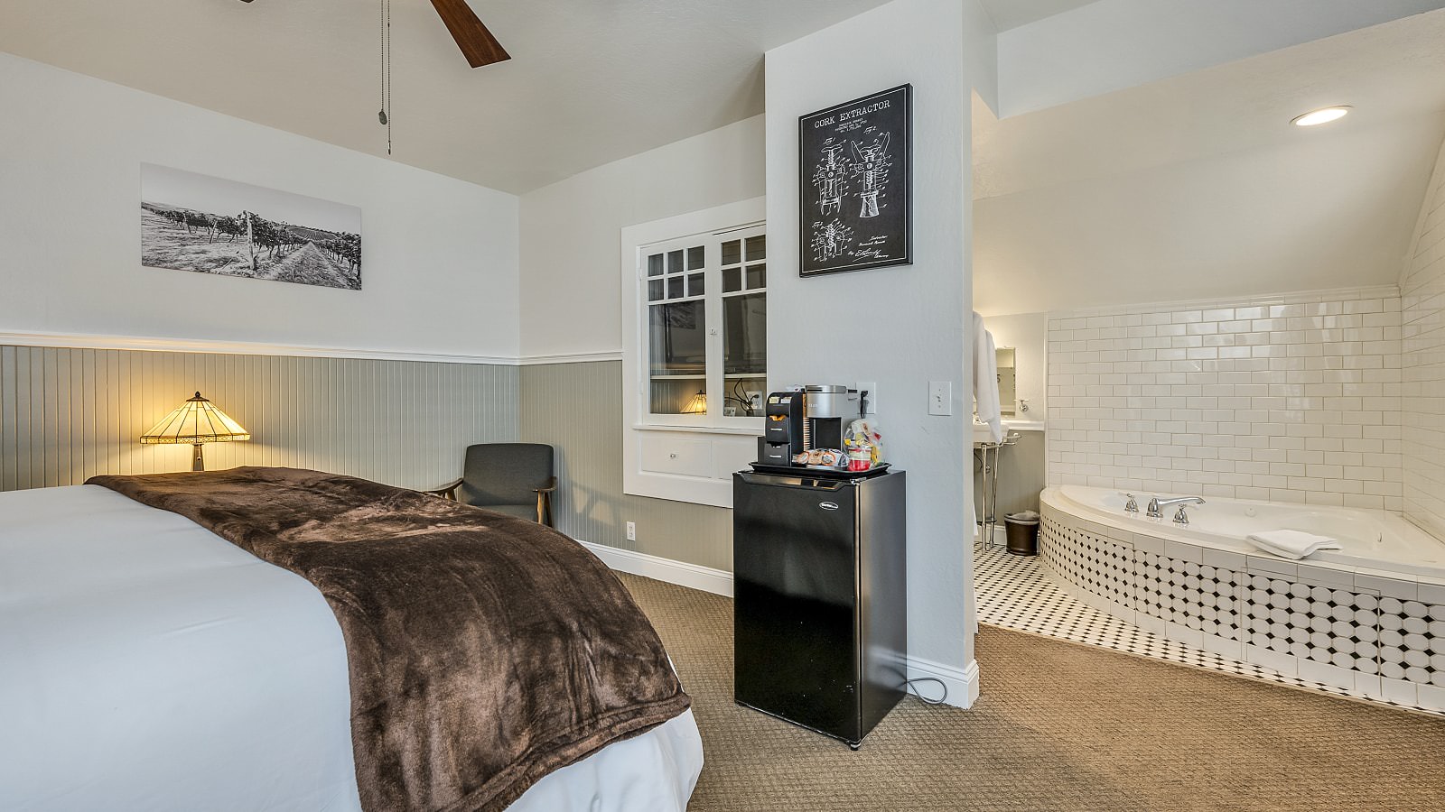 Bedroom with white walls, light gray wainscotting, light colored carpeting, white bedding, black mini fridge, and view into bathroom