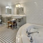 Bathroom with white walls, light gray wainscotting, black and white tiled floor, white toilet, double white pedestal sinks, and soaker tub