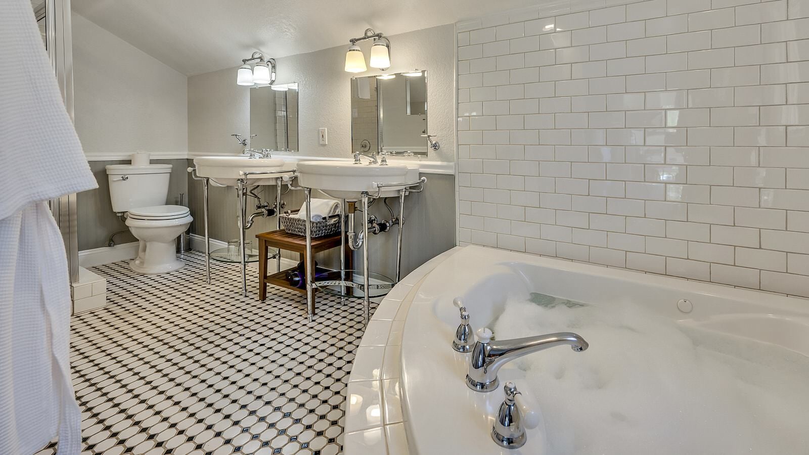 Bathroom with white walls, light gray wainscotting, black and white tiled floor, white toilet, double white pedestal sinks, and soaker tub
