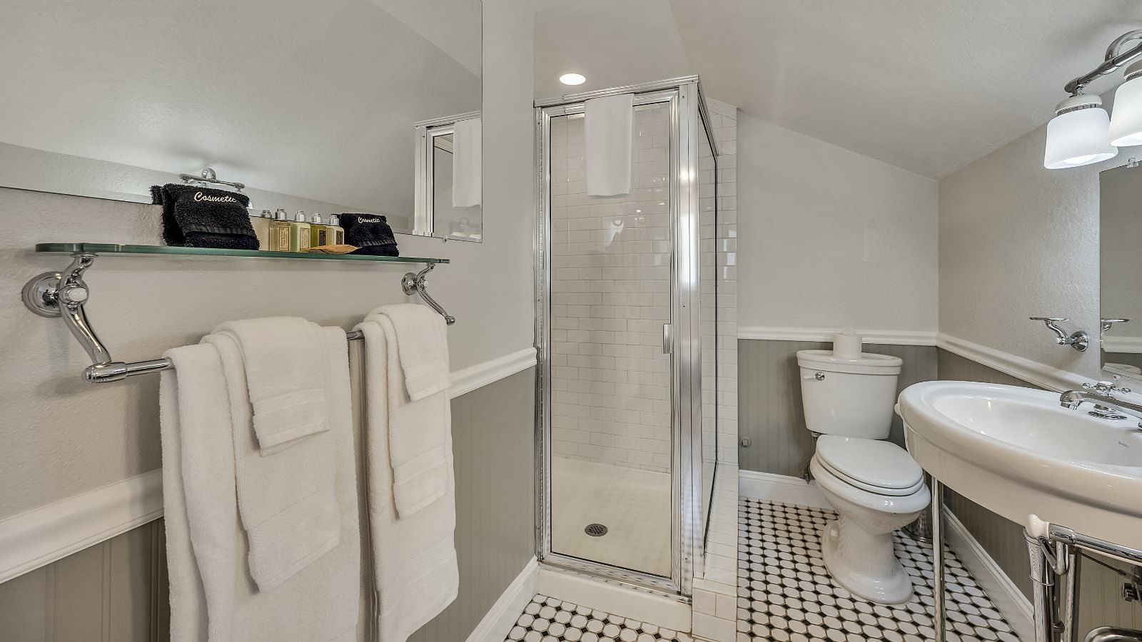 Bathroom with white walls, light gray wainscotting, black and white tiled floor, white toilet, white pedestal sink, and stand up shower