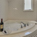 Two wine glasses on glass tray next to bottle of red wine on top of soaker tub surrounded by white tile