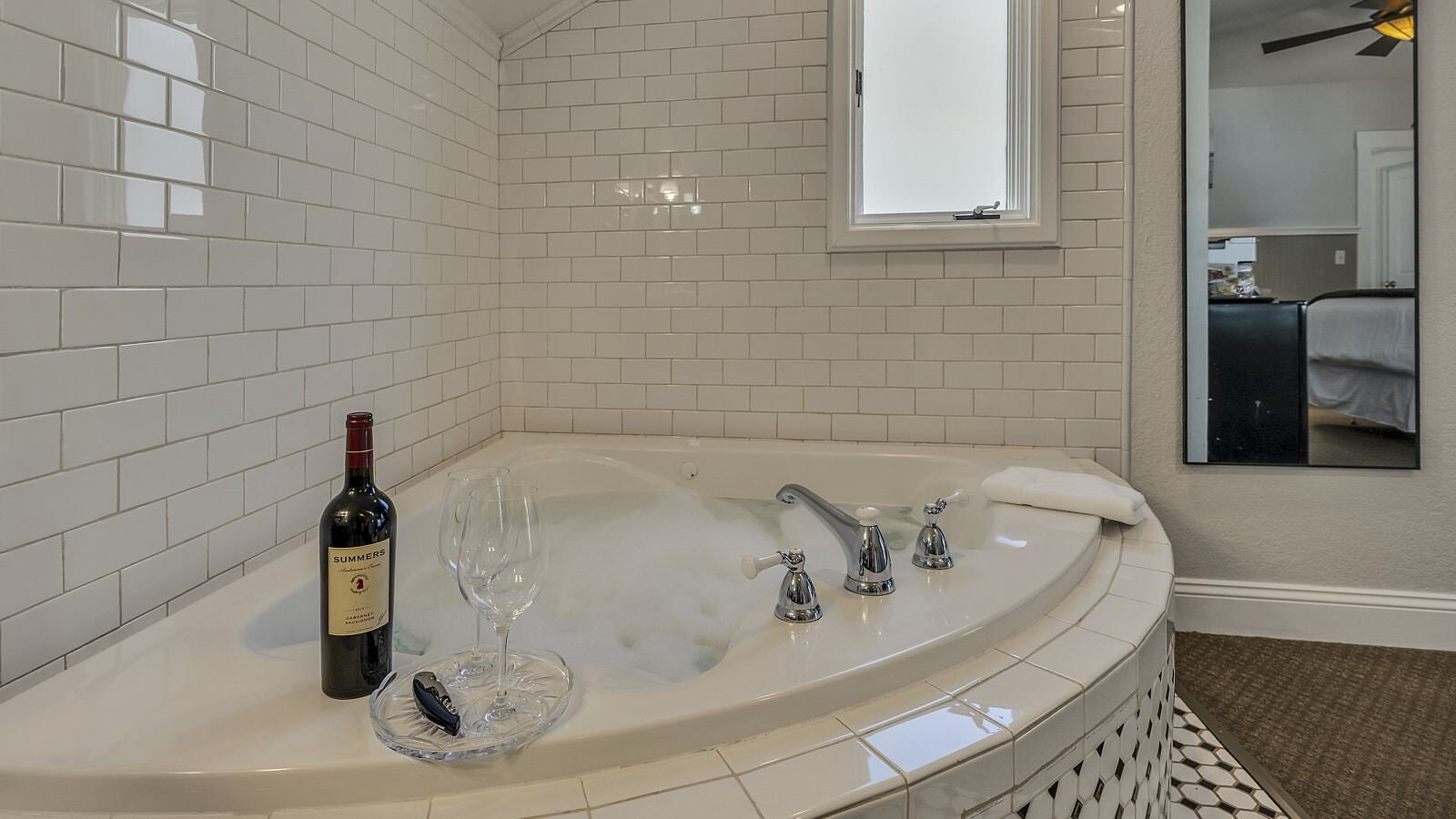 Two wine glasses on glass tray next to bottle of red wine on top of soaker tub surrounded by white tile