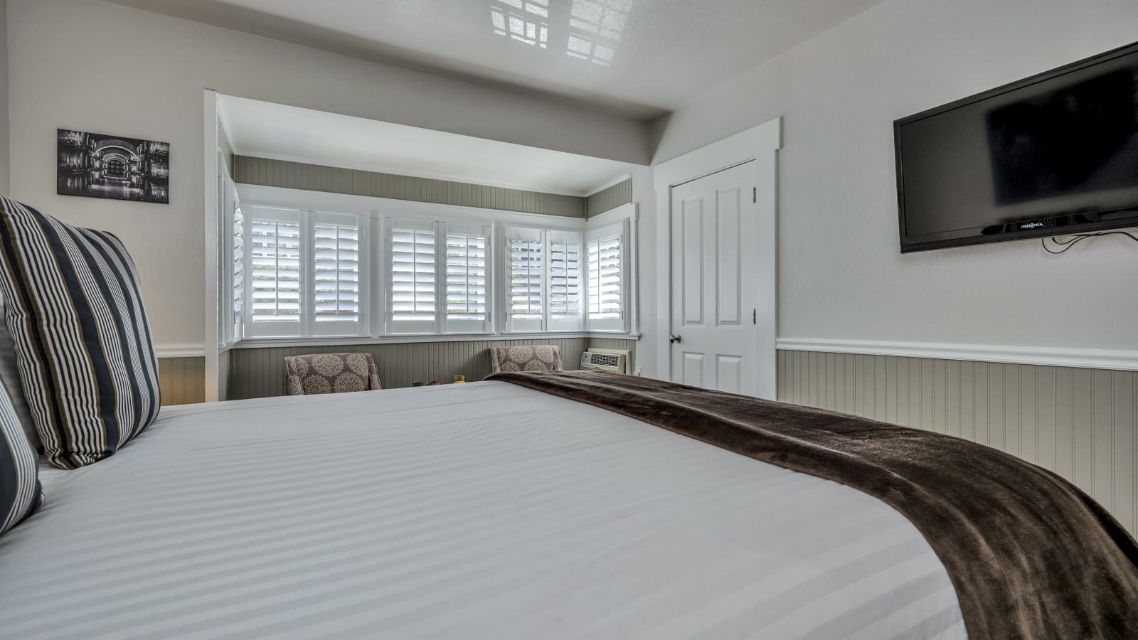 Bedroom with white walls, light gray wainscotting, light colored carpeting, white bedding, and sitting area
