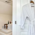 Close up view of white rob hanging on white door with view into bathroom with white-tiled tub