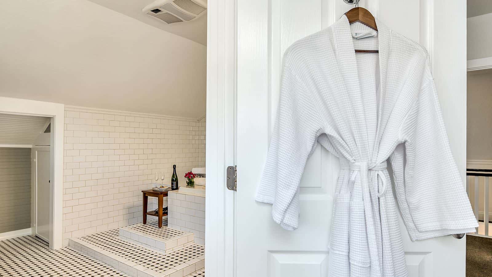Close up view of white rob hanging on white door with view into bathroom with white-tiled tub