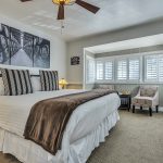 Bedroom with white walls, light gray wainscotting, light colored carpeting, white bedding, dark wooden nightstands, and sitting area