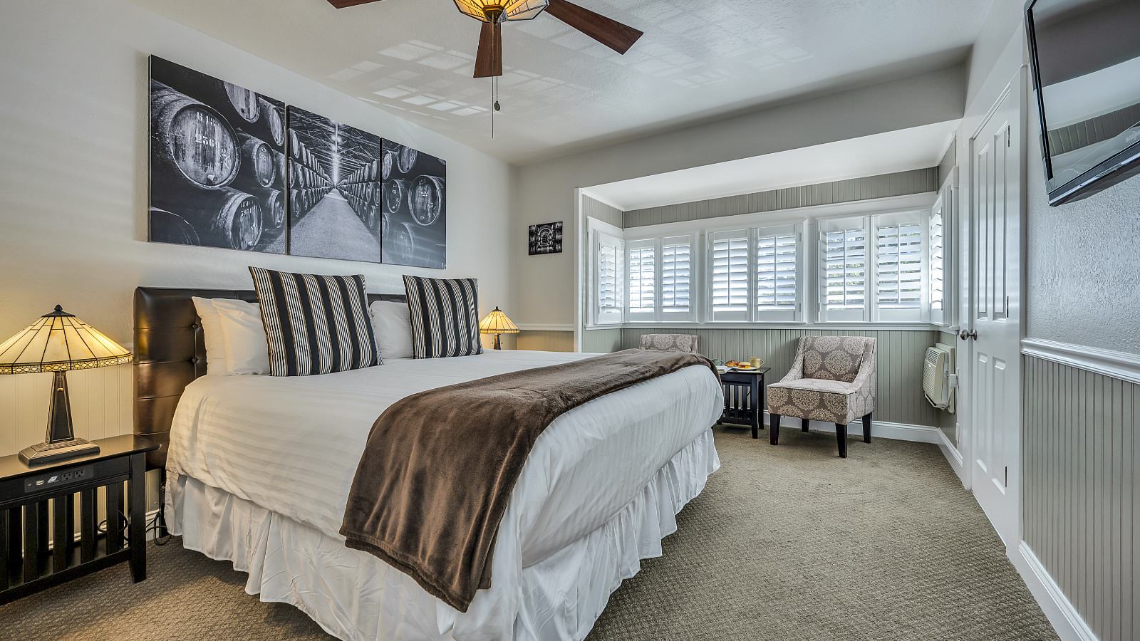 Bedroom with white walls, light gray wainscotting, light colored carpeting, white bedding, dark wooden nightstands, and sitting area