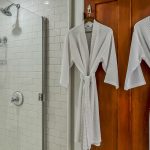 Bathroom with white tiled stand up shower and wooden door with white robes hanging from hooks
