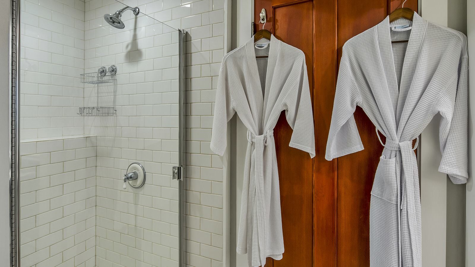 Bathroom with white tiled stand up shower and wooden door with white robes hanging from hooks
