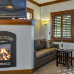 Bedroom with white walls, light colored carpeting, electric fireplace, brown upholstered sofa, and dark wooden end table