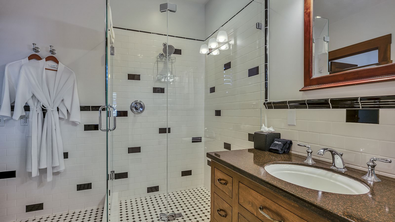 Bathroom with white walls, black and white tiled floor, dark wooden vanity, and black and white tiled shower