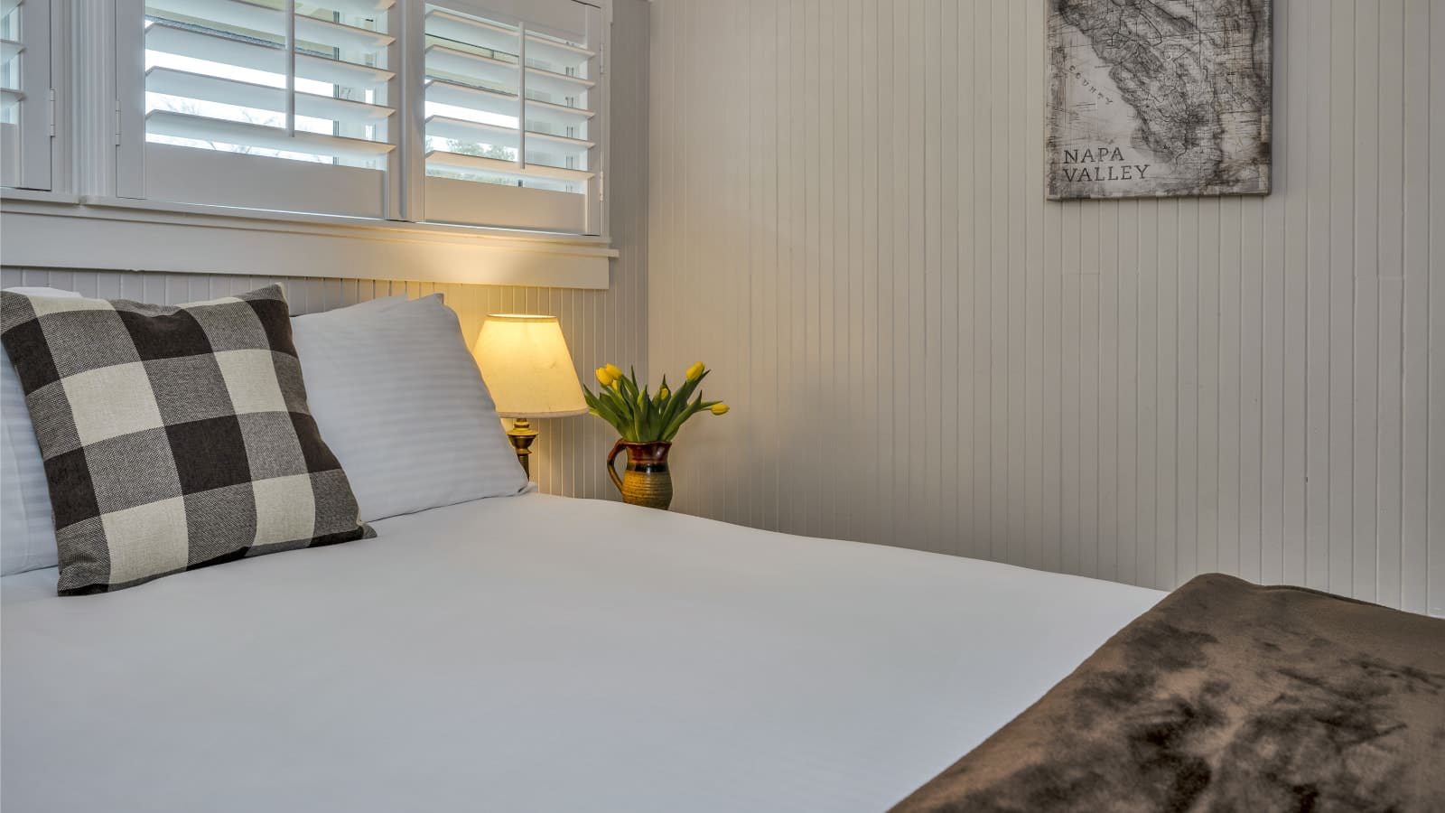 Bedroom with light gray walls, white bedding, and nightstand with lamp and vase of yellow tulips