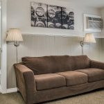 Bedroom with white walls, light gray wainscotting, light colored carpeting, sofa with brown slipcover, and view into bathroom