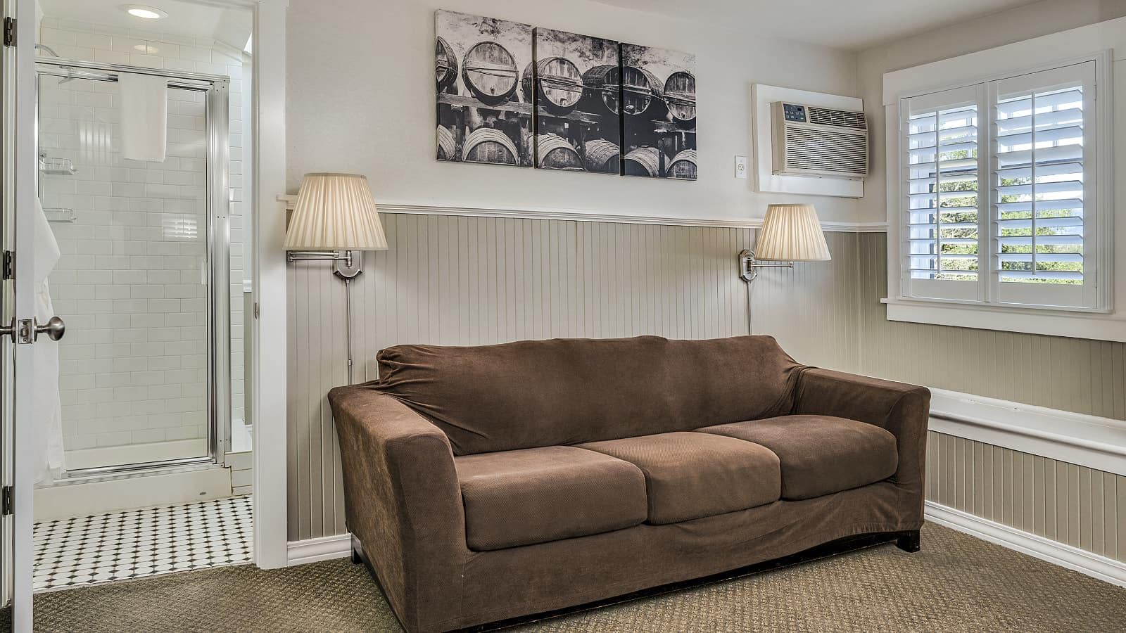Bedroom with white walls, light gray wainscotting, light colored carpeting, sofa with brown slipcover, and view into bathroom