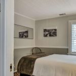 View into bedroom with white walls, light gray wainscotting, floral carpeting, white bedding, and dark wooden chair