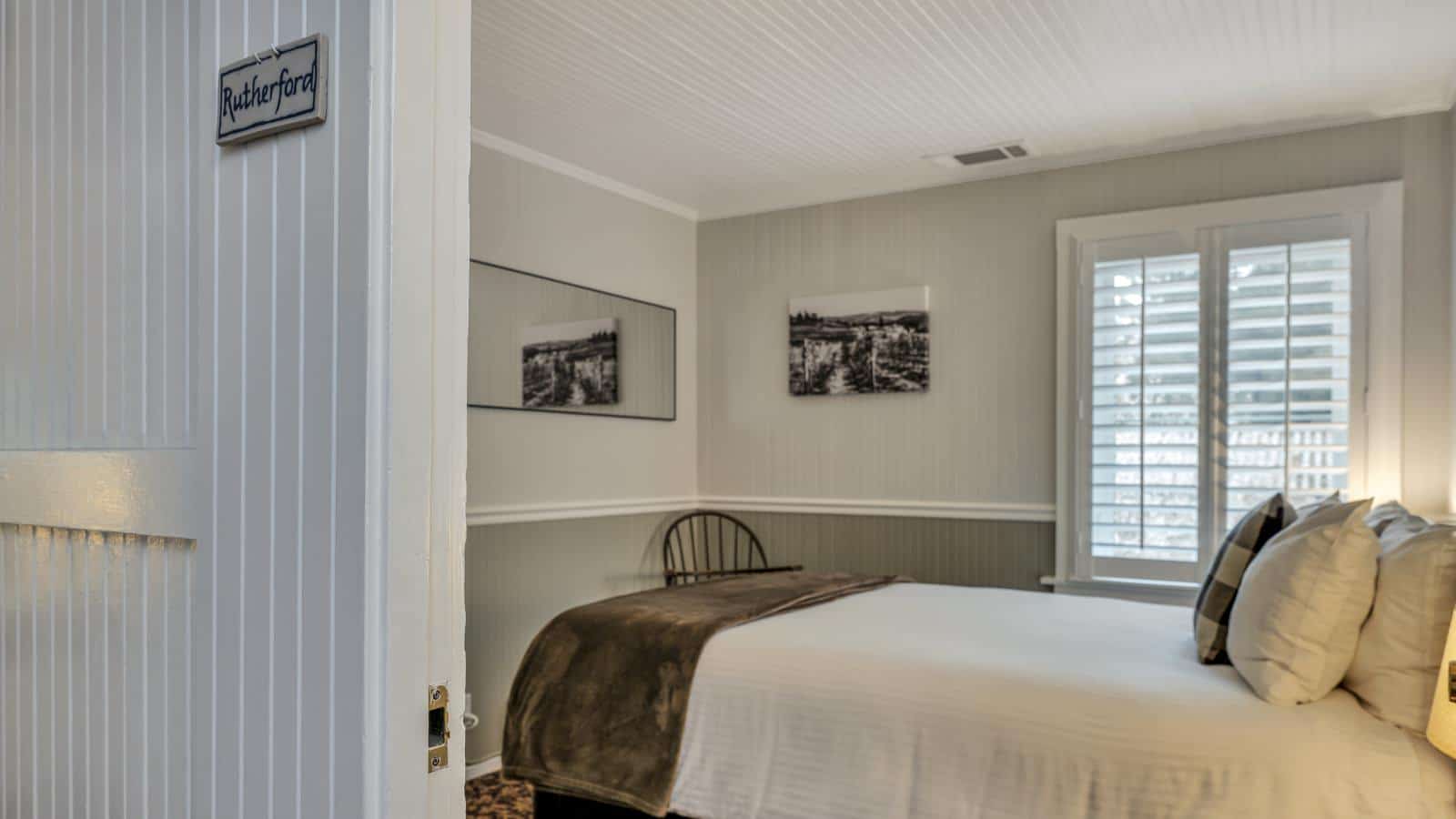 View into bedroom with white walls, light gray wainscotting, floral carpeting, white bedding, and dark wooden chair