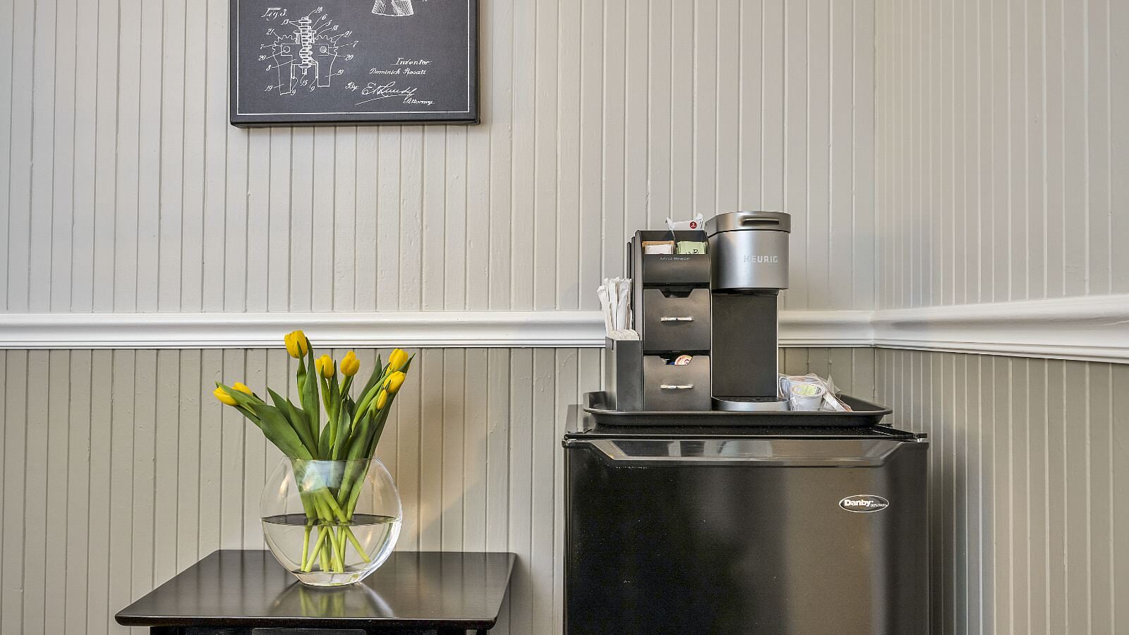 Close up view of black mini fridge with Keurig on top and black table with glass vase with yellow tulips