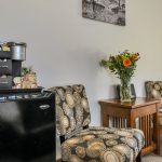 Bedroom sitting area with upholstered chairs, wooden end table, black mini fridge, and Keurig coffee maker