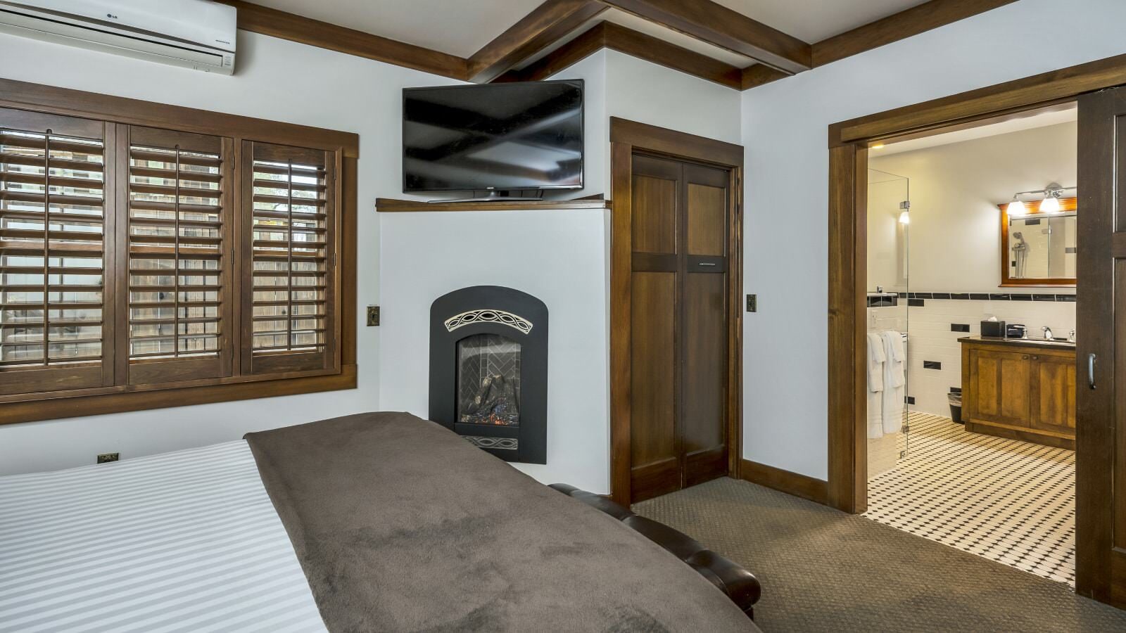 Bedroom with white walls, light colored carpeting, white bedding, leather bed bench, and electric fireplace
