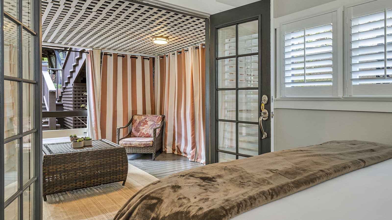 View from bedroom of back wooden deck with brown wicker patio furniture, lattice cover, and white and brown striped curtains