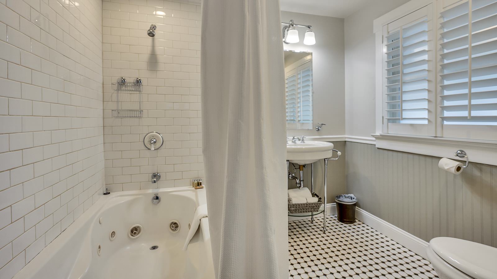 Bathroom with white walls, light gray wainscotting, black and white tiled flooring, white tiled shower and tub combo, and white sink