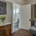 Bedroom with white walls, high light gray wainscotting, floral carpeting, antique wooden dresser, gray upholstered futon, and view into bathroom