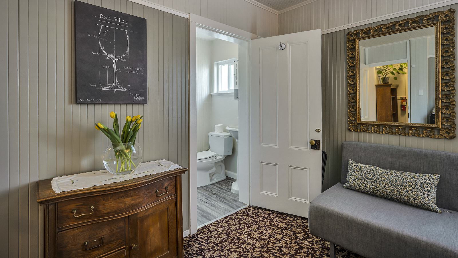 Bedroom with white walls, high light gray wainscotting, floral carpeting, antique wooden dresser, gray upholstered futon, and view into bathroom