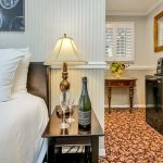 Close up view of bed with leather upholstered head board and white bedding and dark wooden nightstand with lamp, bottle of Champagne, and two Champagne flutes