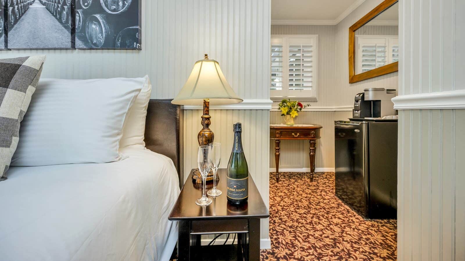 Close up view of bed with leather upholstered head board and white bedding and dark wooden nightstand with lamp, bottle of Champagne, and two Champagne flutes