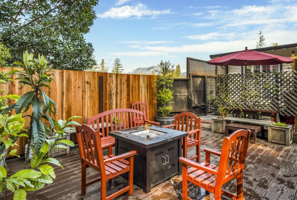 Wooden deck with a firepit surrounded by orange wooden chairs and bench and a patio dining table and chairs with burgundy umbrella