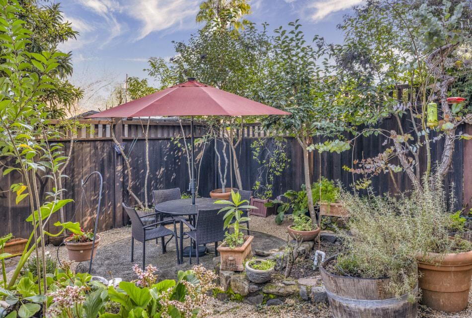 Property's garden with dark wicker patio dining table and chairs with red umbrella and green plants, bushes, and trees