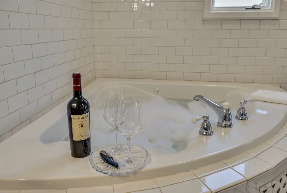 Two wine glasses on glass tray next to bottle of red wine on top of soaker tub surrounded by white tile